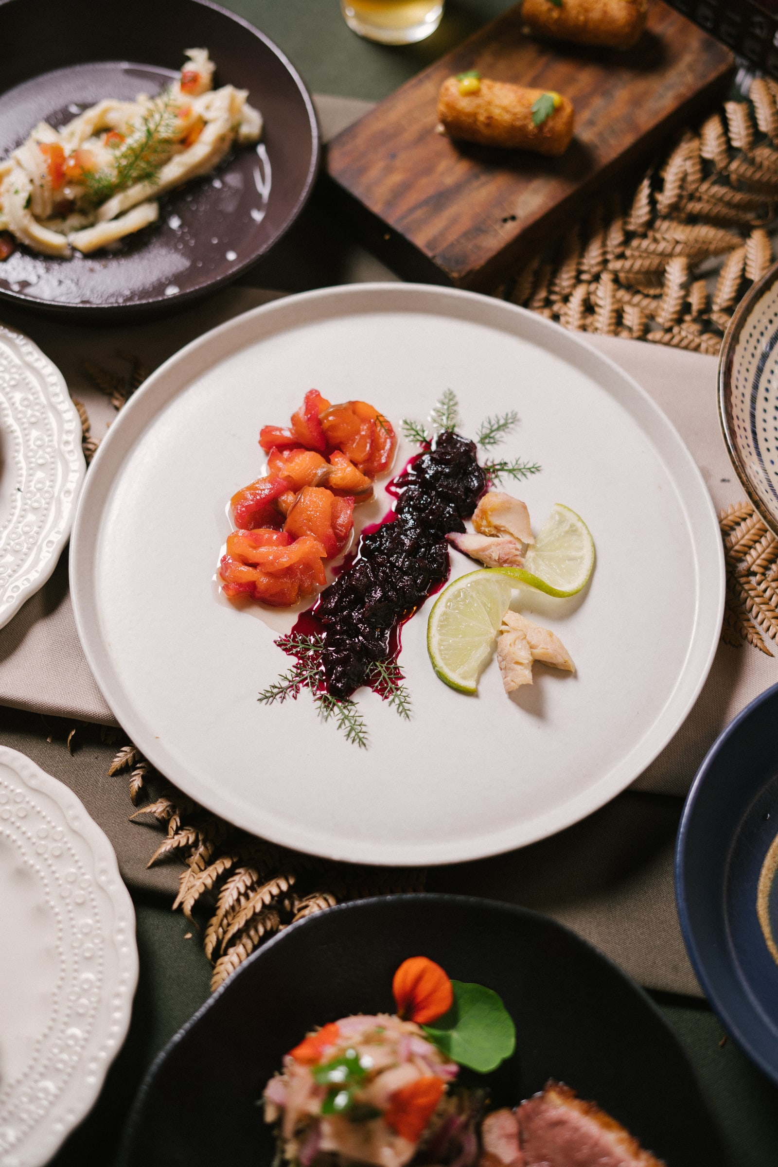 Prato de peixe curado com frutas vermelhas, cercado por outros pratos do Ana Terra